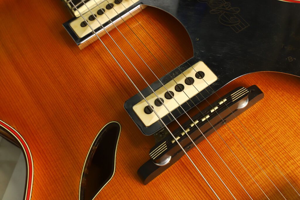 Nahaufnahme des Korpus einer Artur-Lang-Jazzgitarre aus den 1950er Jahren, auf der die Saiten, die Tonabnehmer, der Holzsteg und das F-Loch auf einer polierten, bernsteinfarbenen Oberfläche zu sehen sind. Nahaufnahme des Korpus einer Artur-Lang-Jazzgitarre aus den 1950er Jahren, auf der die Saiten, die Tonabnehmer, der Holzsteg und das F-Loch auf einer polierten, bernsteinfarbenen Oberfläche zu sehen sind.
