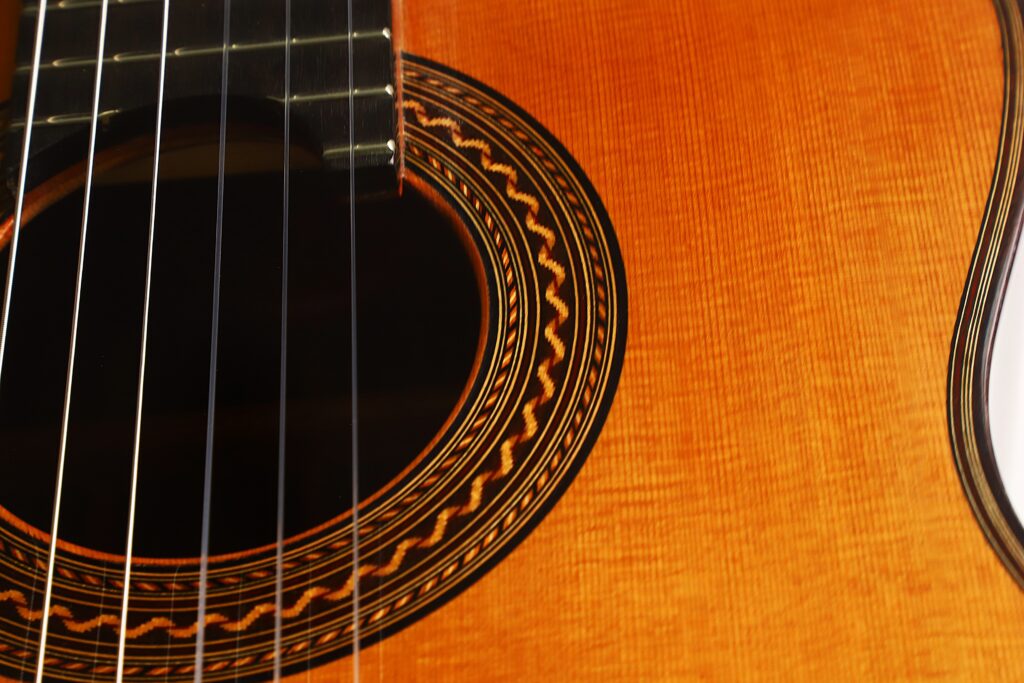 Nahaufnahme des Schalllochs und der Saiten der „Antonius Müller 2012 Doubletop Klassische Gitarre“, die die detaillierte Holzmaserung und das aufwendig gestaltete Rosettenmuster rund um das Schallloch zeigt. Nahaufnahme des Schalllochs und der Saiten der „Antonius Müller 2012 Doubletop Klassische Gitarre“, die die detaillierte Holzmaserung und das aufwendig gestaltete Rosettenmuster rund um das Schallloch zeigt.