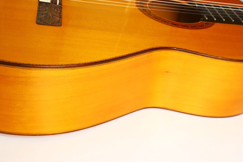 Nahaufnahme des Korpus einer Flamenco-Gitarre von Jeronimo Pena aus dem Jahr 1976, bei der die Holzmaserung und die Saiten hervorgehoben werden, wobei der Fokus auf der glatten, geschwungenen Oberfläche und der glänzenden Lackierung liegt. Nahaufnahme des Korpus einer Flamenco-Gitarre von Jeronimo Pena aus dem Jahr 1976, bei der die Holzmaserung und die Saiten hervorgehoben werden, wobei der Fokus auf der glatten, geschwungenen Oberfläche und der glänzenden Lackierung liegt.