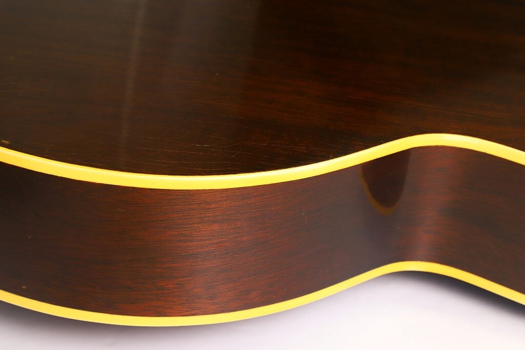 Nahaufnahme der Seite einer Gibson L-48 Akustikgitarre aus dem Jahr 1948, die den braunen Holzkorpus mit glänzender Lackierung und gelber Zierleiste am Rand hervorhebt. Nahaufnahme der Seite einer Gibson L-48 Akustikgitarre aus dem Jahr 1948, die den braunen Holzkorpus mit glänzender Lackierung und gelber Zierleiste am Rand hervorhebt.