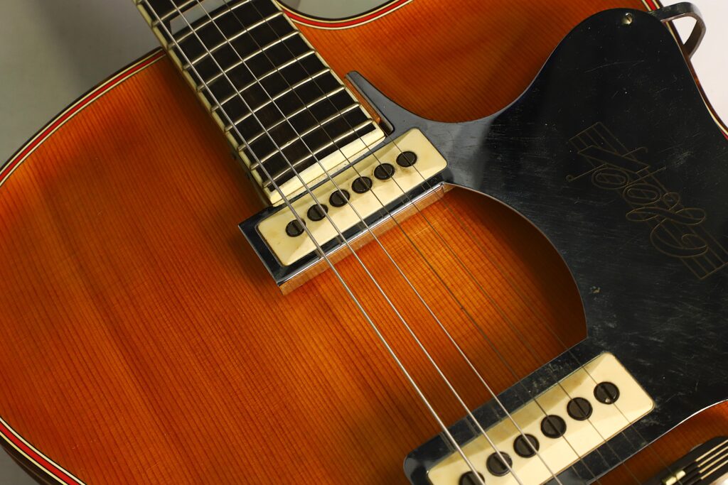 Nahaufnahme einer Artur-Lang-Jazzgitarre aus den 1950er Jahren mit einem Holzkorpus, zwei cremefarbenen Tonabnehmern, einem schwarzen Schlagbrett, sichtbaren Metallsaiten und einer Zierleiste an den Kanten. Nahaufnahme einer Artur-Lang-Jazzgitarre aus den 1950er Jahren mit einem Holzkorpus, zwei cremefarbenen Tonabnehmern, einem schwarzen Schlagbrett, sichtbaren Metallsaiten und einer Zierleiste an den Kanten.