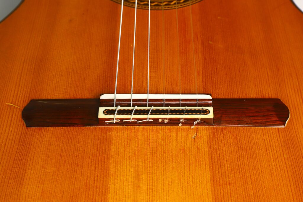 Nahaufnahme des Stegs und der Saiten der „Antonius Müller 2004 Doubletop Klassische Gitarre“, die die feinen Details der Holzmaserung und der Saitenbefestigungen am Holzkorpus hervorhebt. Nahaufnahme des Stegs und der Saiten der „Antonius Müller 2004 Doubletop Klassische Gitarre“, die die feinen Details der Holzmaserung und der Saitenbefestigungen am Holzkorpus hervorhebt.
