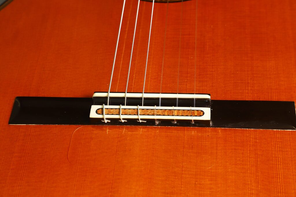 Nahaufnahme des Sattels der „Antonius Müller 2012 Doubletop Klassische Gitarre“ mit sechs aufgezogenen Saiten, wobei der Holzkorpus, der Sattel und die filigranen Saitenknoten vor dem Hintergrund der polierten Oberfläche besonders hervorgehoben werden. Nahaufnahme des Sattels der „Antonius Müller 2012 Doubletop Klassische Gitarre“ mit sechs aufgezogenen Saiten, wobei der Holzkorpus, der Sattel und die filigranen Saitenknoten vor dem Hintergrund der polierten Oberfläche besonders hervorgehoben werden.