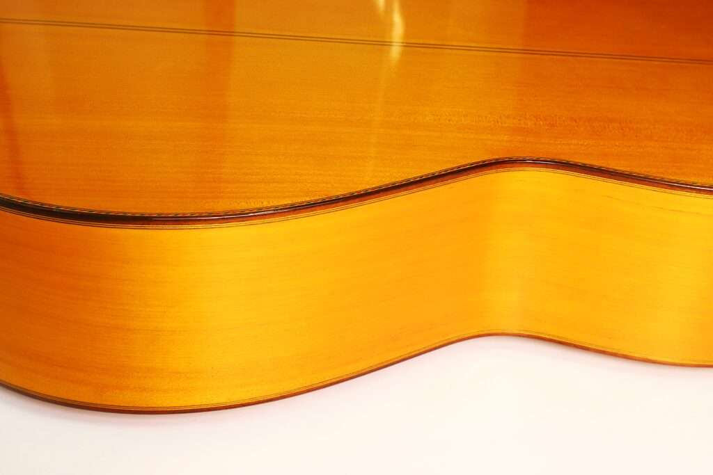 Nahaufnahme der Seite und Rückseite einer glänzenden Flamenco-Gitarre von Jeronimo Pena aus dem Jahr 1976 aus hellem Holz vor weißem Hintergrund. Die Rundungen und die glatte Oberfläche des Instruments werden besonders hervorgehoben. Nahaufnahme der Seite und Rückseite einer glänzenden Flamenco-Gitarre von Jeronimo Pena aus dem Jahr 1976 aus hellem Holz vor weißem Hintergrund. Die Rundungen und die glatte Oberfläche des Instruments werden besonders hervorgehoben.