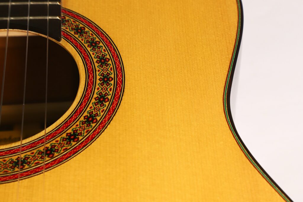 Nahaufnahme des Korpus der „Gerhard Arns 2026 Smallman Klassische Gitarre“, die einen Teil des Schalllochs mit einer dekorativen rot-schwarzen Rosette sowie die Maserung der Gitarrendecke zeigt. Nahaufnahme des Korpus der „Gerhard Arns 2026 Smallman Klassische Gitarre“, die einen Teil des Schalllochs mit einer dekorativen rot-schwarzen Rosette sowie die Maserung der Gitarrendecke zeigt.