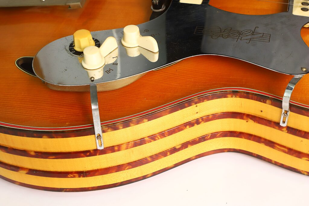 Nahaufnahme der Artur Lang Jazz-Gitarre aus den 1950er Jahren, bei der die Regler, das Schlagbrett mit Beschriftung sowie der mehrschichtige, gestreifte Holzkorpus mit Zierkanten und Metallhalterungen besonders hervorgehoben werden. Nahaufnahme der Artur Lang Jazz-Gitarre aus den 1950er Jahren, bei der die Regler, das Schlagbrett mit Beschriftung sowie der mehrschichtige, gestreifte Holzkorpus mit Zierkanten und Metallhalterungen besonders hervorgehoben werden.