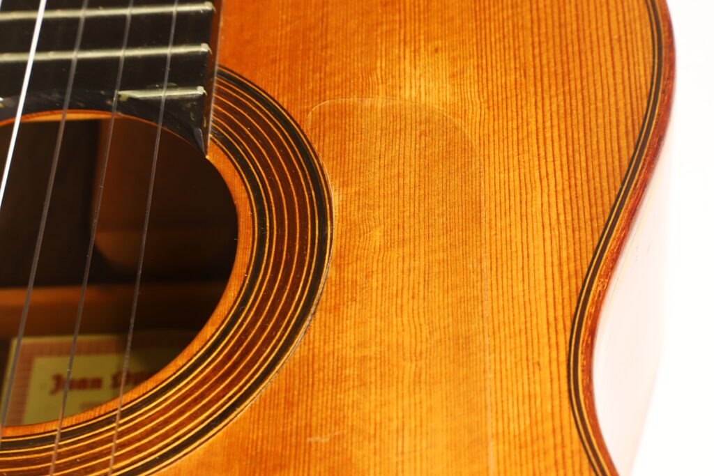 Nahaufnahme des Holzkörpers der Flamenco-Gitarre „Juan Lopez Aguilarte 1984 Negra“, die die Saiten, das mit schwarzen und weißen konzentrischen Ringen verzierte Schallloch sowie die filigrane Maserung des Holzes zeigt. Das Bild verdeutlicht die außergewöhnliche Handwerkskunst und die reichhaltige Textur dieser Flamenco-Gitarre. Nahaufnahme des Holzkörpers der Flamenco-Gitarre „Juan Lopez Aguilarte 1984 Negra“, die die Saiten, das mit schwarzen und weißen konzentrischen Ringen verzierte Schallloch sowie die filigrane Maserung des Holzes zeigt. Das Bild verdeutlicht die außergewöhnliche Handwerkskunst und die reichhaltige Textur dieser Flamenco-Gitarre.