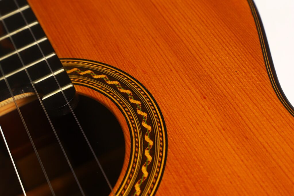 Nahaufnahme der „Antonius Müller 2004 Doubletop Klassische Gitarre“, die einen Teil der Saiten, das Schallloch und die dekorative Rosette auf dem Holzkorpus zeigt. Das Bild verdeutlicht die Textur und die handwerkliche Qualität dieser meisterhaft gefertigten klassischen Gitarre. Nahaufnahme der „Antonius Müller 2004 Doubletop Klassische Gitarre“, die einen Teil der Saiten, das Schallloch und die dekorative Rosette auf dem Holzkorpus zeigt. Das Bild verdeutlicht die Textur und die handwerkliche Qualität dieser meisterhaft gefertigten klassischen Gitarre.