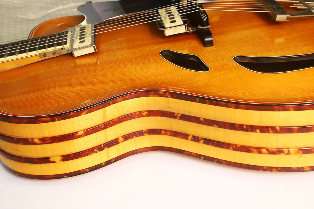 Nahaufnahme des Korpus einer Artur-Lang-Jazzgitarre aus den 1950er Jahren mit drei wellenförmigen, geschichteten Kanten und zwei f-förmigen Schalllöchern. Diese Gitarre verfügt über eine Hochglanzlackierung und ein markantes Streifenmuster an den Seiten. Nahaufnahme des Korpus einer Artur-Lang-Jazzgitarre aus den 1950er Jahren mit drei wellenförmigen, geschichteten Kanten und zwei f-förmigen Schalllöchern. Diese Gitarre verfügt über eine Hochglanzlackierung und ein markantes Streifenmuster an den Seiten.