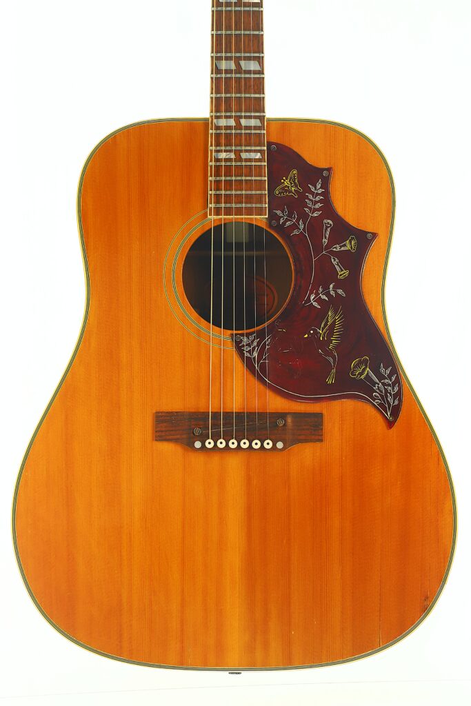 Gibson Hummingbird 1968 Akustikgitarre mit naturbelassener Holzoberfläche und einem dekorativen Schlagbrett mit Blumenmustern und einem Kolibri-Motiv, fotografiert vor weißem Hintergrund. Gibson Hummingbird 1968 Akustikgitarre mit naturbelassener Holzoberfläche und einem dekorativen Schlagbrett mit Blumenmustern und einem Kolibri-Motiv, fotografiert vor weißem Hintergrund.