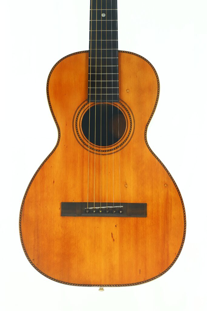 Vorderansicht der Washburn 1900 Parlor Akustikgitarre mit Holzkorpus, sechs Saiten, dunklem Griffbrett und dekorativen Einlagen rund um das Schallloch und an den Kanten, fotografiert vor weißem Hintergrund. Vorderansicht der Washburn 1900 Parlor Akustikgitarre mit Holzkorpus, sechs Saiten, dunklem Griffbrett und dekorativen Einlagen rund um das Schallloch und an den Kanten, fotografiert vor weißem Hintergrund.
