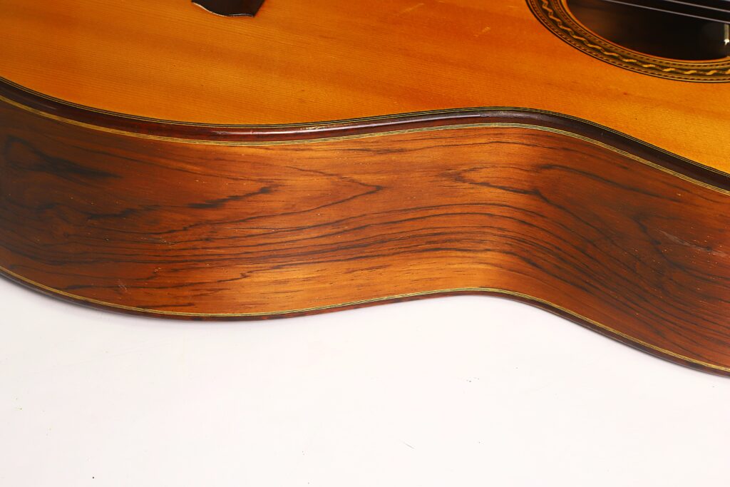 Nahaufnahme der gewölbten Holzseite einer „Antonius Müller 2004 Doubletop Klassische Gitarre“, die die Holzmaserung und die glänzende Lackierung zeigt, wobei ein Teil der Decke und die Zierrosette der Gitarre zu sehen sind. Nahaufnahme der gewölbten Holzseite einer „Antonius Müller 2004 Doubletop Klassische Gitarre“, die die Holzmaserung und die glänzende Lackierung zeigt, wobei ein Teil der Decke und die Zierrosette der Gitarre zu sehen sind.
