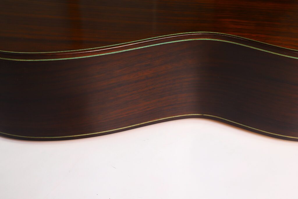 Nahaufnahme der Seite der „Gerhard Arns 2026 Smallman Klassische Gitarre“, die den dunklen Holzkorpus mit hellen Zierleisten zeigt und die eleganten Rundungen des Instruments vor einem weißen Hintergrund hervorhebt. Nahaufnahme der Seite der „Gerhard Arns 2026 Smallman Klassische Gitarre“, die den dunklen Holzkorpus mit hellen Zierleisten zeigt und die eleganten Rundungen des Instruments vor einem weißen Hintergrund hervorhebt.