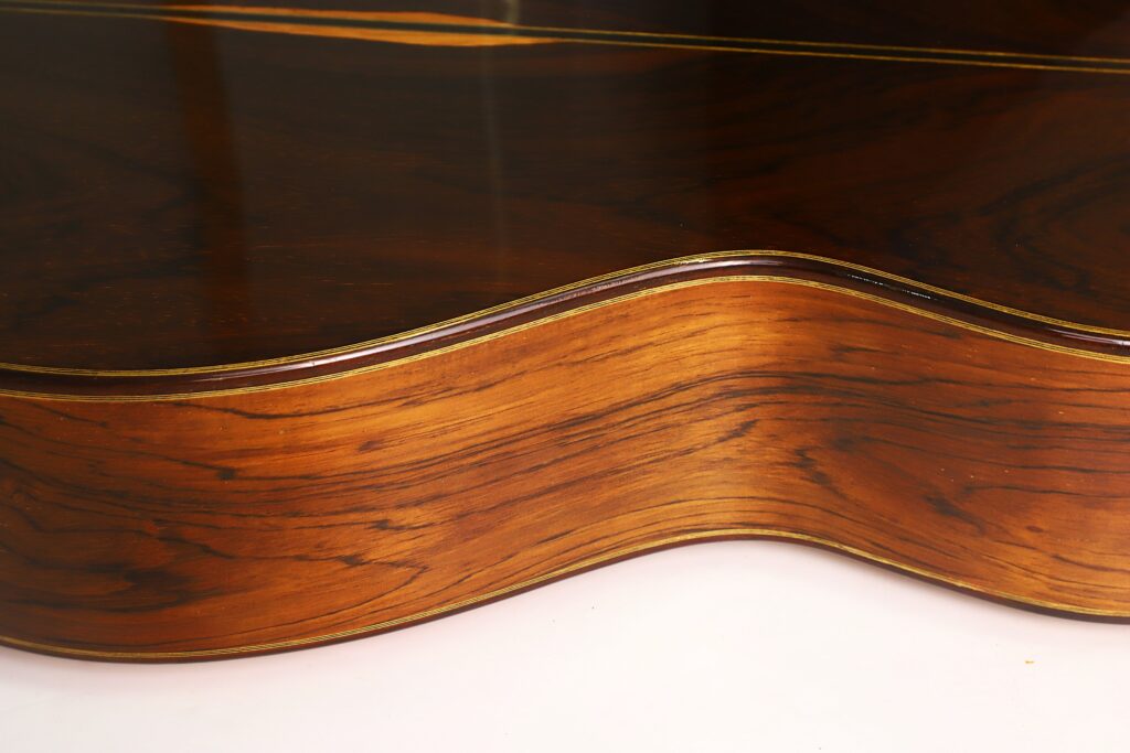 Nahaufnahme der Seiten- und Rückseite der „Antonius Müller 2004 Doubletop Klassische Gitarre“, die die glatte, polierte Holzmaserung und die sanften Rundungen vor weißem Hintergrund hervorhebt. Nahaufnahme der Seiten- und Rückseite der „Antonius Müller 2004 Doubletop Klassische Gitarre“, die die glatte, polierte Holzmaserung und die sanften Rundungen vor weißem Hintergrund hervorhebt.