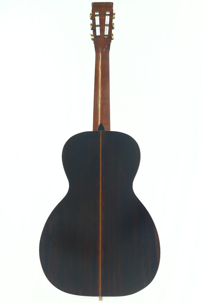 Die Washburn 1900 Parlor-Akustikgitarre ist von hinten fotografiert und zeigt ihren dunklen Holzkorpus mit einer dekorativen Mittelnaht sowie einen helleren Holzhals, der mit Stimmwirbeln versehen ist. Die Washburn 1900 Parlor-Akustikgitarre ist von hinten fotografiert und zeigt ihren dunklen Holzkorpus mit einer dekorativen Mittelnaht sowie einen helleren Holzhals, der mit Stimmwirbeln versehen ist.
