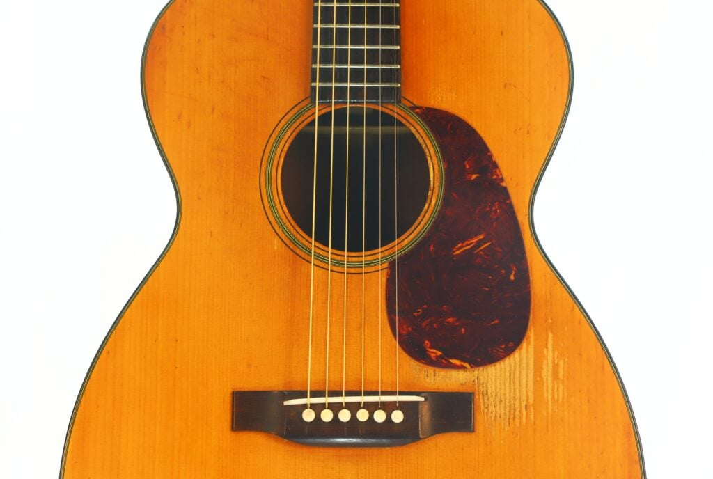 Nahaufnahme einer Martin 00-18 Akustikgitarre aus dem Jahr 1953, bei der die Saiten, das Schallloch, das Schlagbrett und der Steg vor einem weißen Hintergrund hervorgehoben sind. Die Gitarre verfügt über eine warme, natürliche Holzoberfläche. Nahaufnahme einer Martin 00-18 Akustikgitarre aus dem Jahr 1953, bei der die Saiten, das Schallloch, das Schlagbrett und der Steg vor einem weißen Hintergrund hervorgehoben sind. Die Gitarre verfügt über eine warme, natürliche Holzoberfläche.