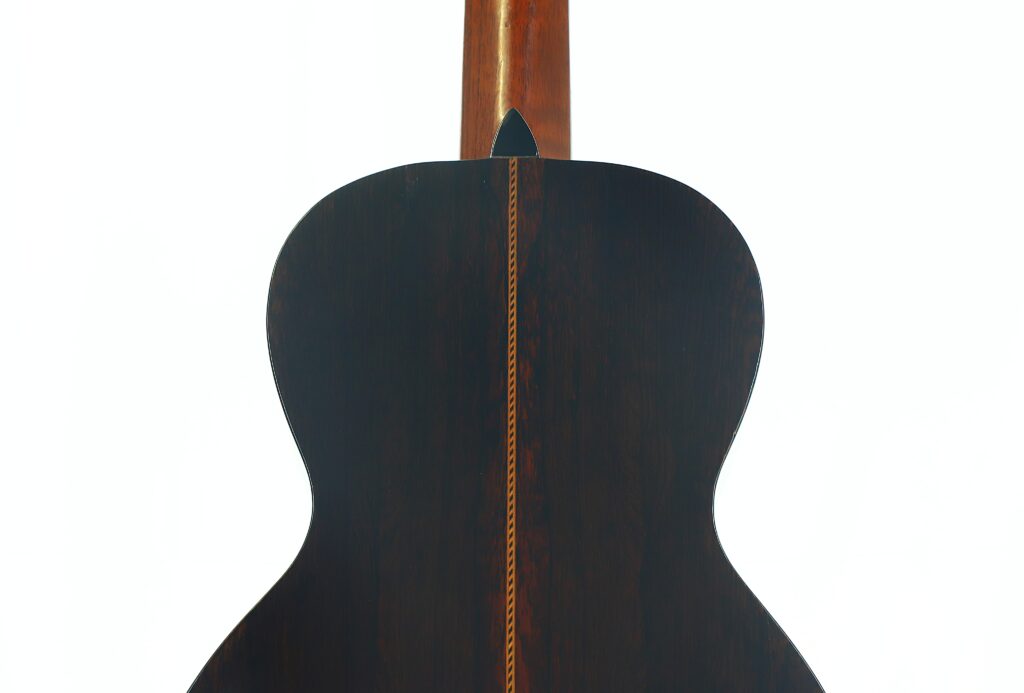 Die Rückseite der Washburn 1900 Parlor-Akustikgitarre, gefertigt aus edlem, dunklem Holz und verziert mit einem dekorativen Einlegeband, das vertikal in der Mitte verläuft. Der Hals der Gitarre ist oben vor einem weißen Hintergrund teilweise zu sehen. Die Rückseite der Washburn 1900 Parlor-Akustikgitarre, gefertigt aus edlem, dunklem Holz und verziert mit einem dekorativen Einlegeband, das vertikal in der Mitte verläuft. Der Hals der Gitarre ist oben vor einem weißen Hintergrund teilweise zu sehen.
