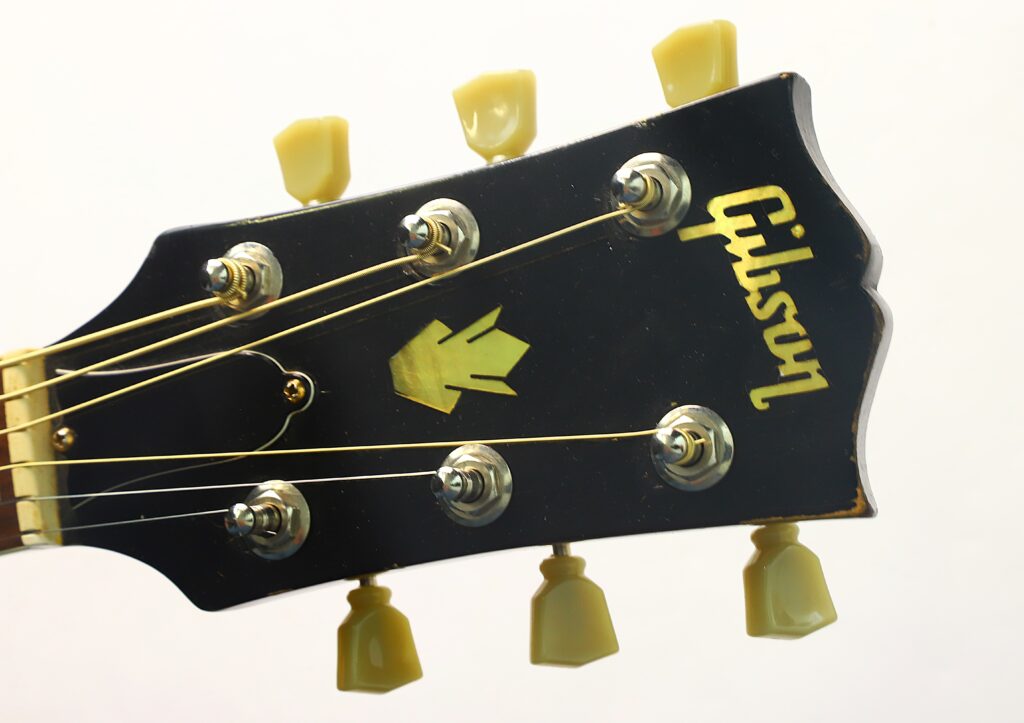 Nahaufnahme des Kopfes einer schwarzen Gibson Hummingbird Akustikgitarre aus dem Jahr 1968 mit goldenem Gibson-Logo und sechs Stimmwirbeln vor einem schlichten weißen Hintergrund. Nahaufnahme des Kopfes einer schwarzen Gibson Hummingbird Akustikgitarre aus dem Jahr 1968 mit goldenem Gibson-Logo und sechs Stimmwirbeln vor einem schlichten weißen Hintergrund.