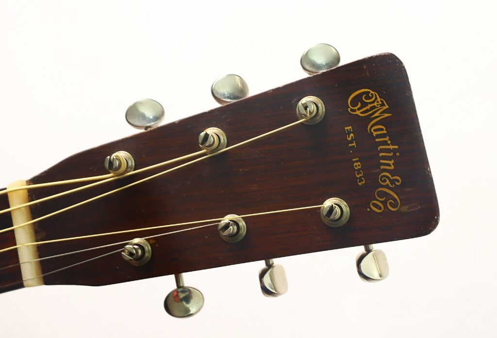 Nahaufnahme des Kopfes einer Martin 00-18 Akustikgitarre aus dem Jahr 1953 mit sechs Stimmwirbeln, Saiten und einem goldenen „Martin & Co.“-Logo mit der Aufschrift „Est. 1833“ auf einem Holzhintergrund. Nahaufnahme des Kopfes einer Martin 00-18 Akustikgitarre aus dem Jahr 1953 mit sechs Stimmwirbeln, Saiten und einem goldenen „Martin & Co.“-Logo mit der Aufschrift „Est. 1833“ auf einem Holzhintergrund.