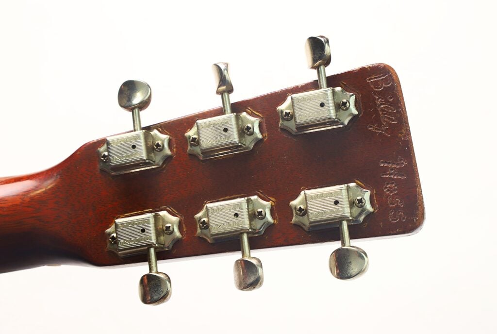 Nahaufnahme der Rückseite des Kopfes einer Martin 00-18 Akustikgitarre aus dem Jahr 1953, auf der sechs Metallstimmwirbel und eine Gravur im Holz zu sehen sind; fotografiert vor weißem Hintergrund. Nahaufnahme der Rückseite des Kopfes einer Martin 00-18 Akustikgitarre aus dem Jahr 1953, auf der sechs Metallstimmwirbel und eine Gravur im Holz zu sehen sind; fotografiert vor weißem Hintergrund.