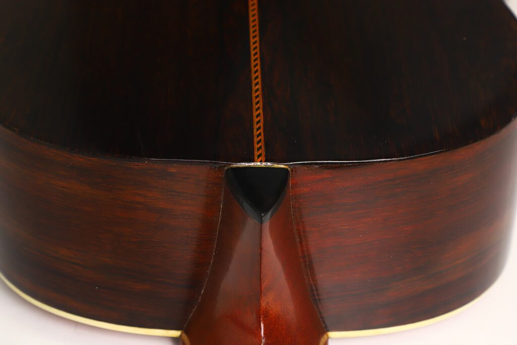 Nahaufnahme der Rückseite und der Seite der Washburn 1900 Parlor Akustikgitarre, die dunkel poliertes Holz, eine Zierintarsie in der Mitte und eine cremefarbene Einfassung an den Kanten zeigt. Nahaufnahme der Rückseite und der Seite der Washburn 1900 Parlor Akustikgitarre, die dunkel poliertes Holz, eine Zierintarsie in der Mitte und eine cremefarbene Einfassung an den Kanten zeigt.