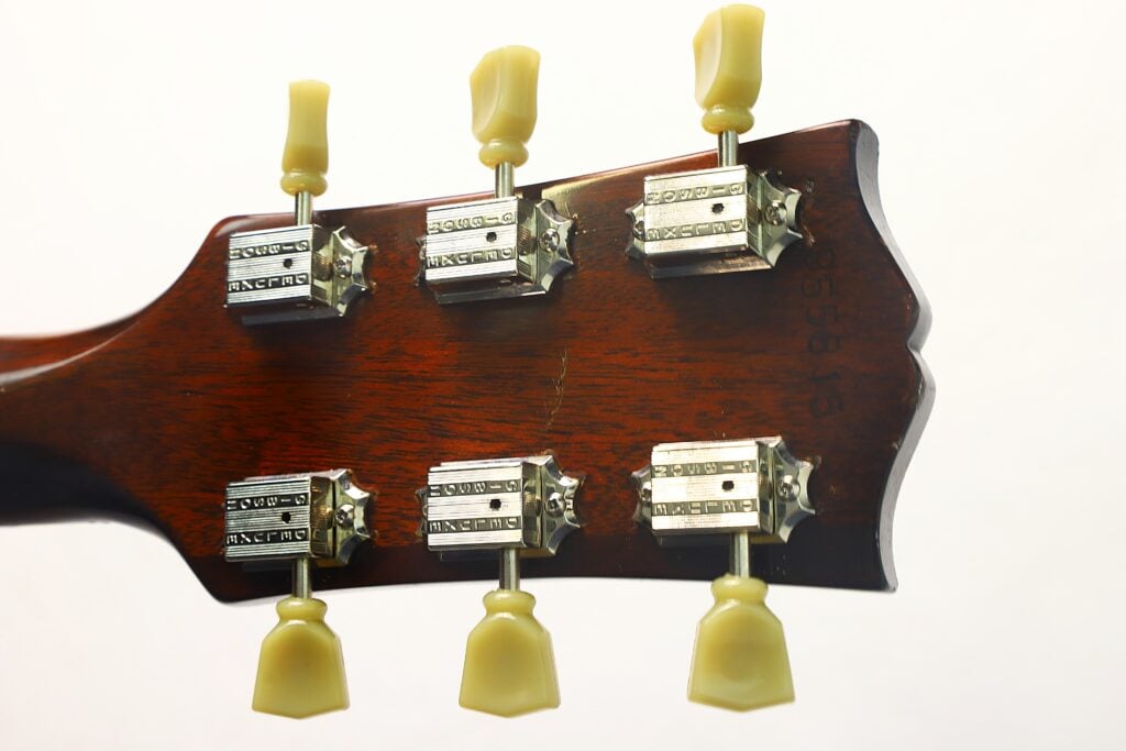 Nahaufnahme der Rückseite des Kopfes einer Gibson Hummingbird Akustikgitarre aus dem Jahr 1968, die sechs Stimmwirbel im Vintage-Stil mit cremefarbenen Knöpfen und metallischen Gehäusen vor einem schlichten Hintergrund zeigt. Nahaufnahme der Rückseite des Kopfes einer Gibson Hummingbird Akustikgitarre aus dem Jahr 1968, die sechs Stimmwirbel im Vintage-Stil mit cremefarbenen Knöpfen und metallischen Gehäusen vor einem schlichten Hintergrund zeigt.