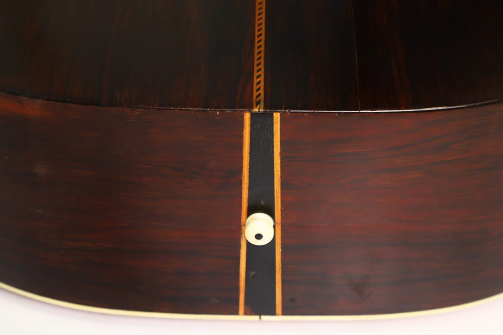 Nahaufnahme der Seiten- und Unterkante der Washburn 1900 Parlor-Akustikgitarre, die eine dunkle Holzmaserung, eine Zierintarsie und einen kleinen weißen Gurtknopf zeigt. Nahaufnahme der Seiten- und Unterkante der Washburn 1900 Parlor-Akustikgitarre, die eine dunkle Holzmaserung, eine Zierintarsie und einen kleinen weißen Gurtknopf zeigt.