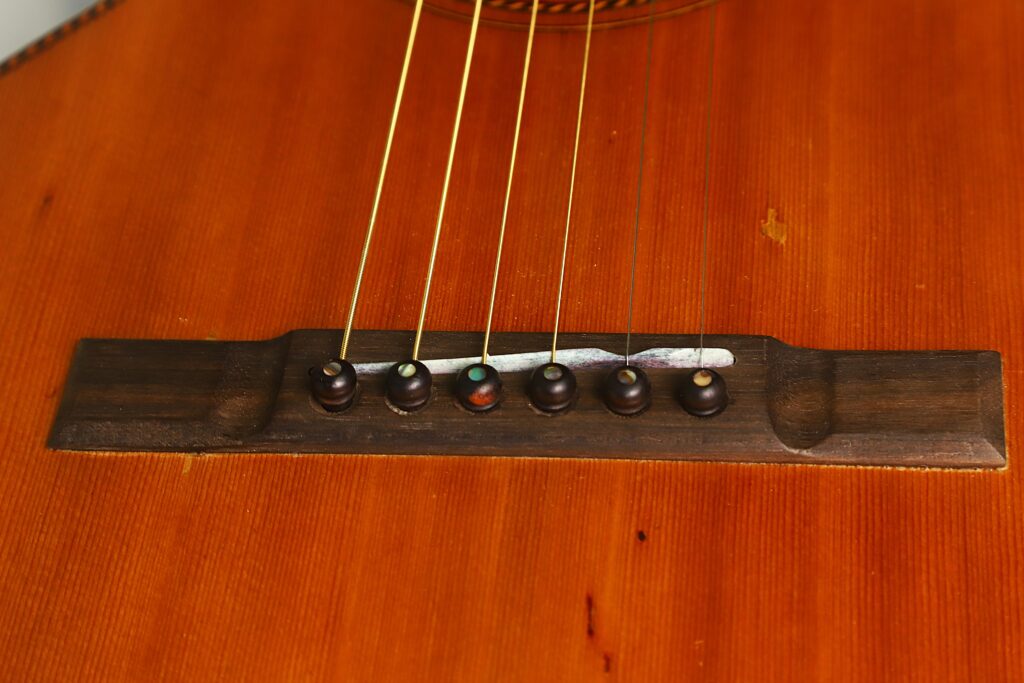 Nahaufnahme des Stegs der Washburn 1900 Parlor Akustikgitarre, auf der die sechs Saiten zu sehen sind, die mit dunklen, runden Stegstiften auf der Holzdecke befestigt sind. Nahaufnahme des Stegs der Washburn 1900 Parlor Akustikgitarre, auf der die sechs Saiten zu sehen sind, die mit dunklen, runden Stegstiften auf der Holzdecke befestigt sind.