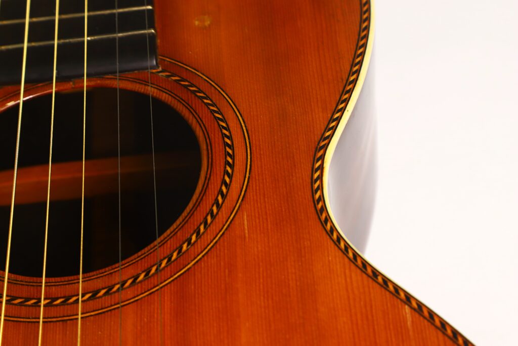 Nahaufnahme des Schalllochs, der Saiten und eines Teils des Korpus der Washburn 1900 Parlor Akustikgitarre, wobei die Holzmaserung und die detaillierten Einlegearbeiten am Rand besonders hervorgehoben werden. Nahaufnahme des Schalllochs, der Saiten und eines Teils des Korpus der Washburn 1900 Parlor Akustikgitarre, wobei die Holzmaserung und die detaillierten Einlegearbeiten am Rand besonders hervorgehoben werden.