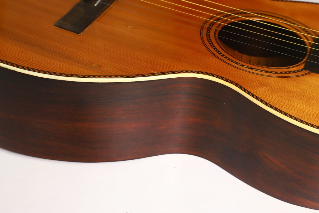 Nahaufnahme des Korpus der Washburn 1900 Parlor Akustikgitarre, die die Holzmaserung, die Saiten und das Schallloch hervorhebt, mit einer Zierkante und einer glatten, geschwungenen Oberfläche vor einem weißen Hintergrund. Nahaufnahme des Korpus der Washburn 1900 Parlor Akustikgitarre, die die Holzmaserung, die Saiten und das Schallloch hervorhebt, mit einer Zierkante und einer glatten, geschwungenen Oberfläche vor einem weißen Hintergrund.