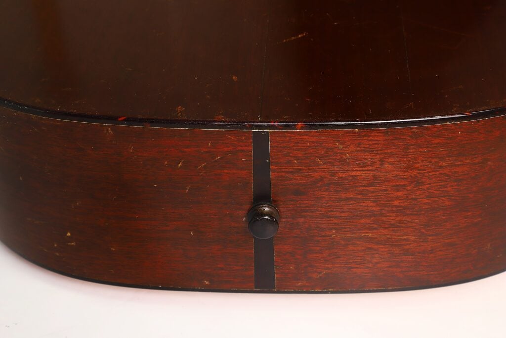 Nahaufnahme einer polierten, dunklen Martin 00-18 Akustikgitarre aus dem Jahr 1953 mit einem schwarzen Drehknopf in der Mitte und einem dünnen schwarzen vertikalen Streifen dahinter, vor einem weißen Hintergrund. Nahaufnahme einer polierten, dunklen Martin 00-18 Akustikgitarre aus dem Jahr 1953 mit einem schwarzen Drehknopf in der Mitte und einem dünnen schwarzen vertikalen Streifen dahinter, vor einem weißen Hintergrund.