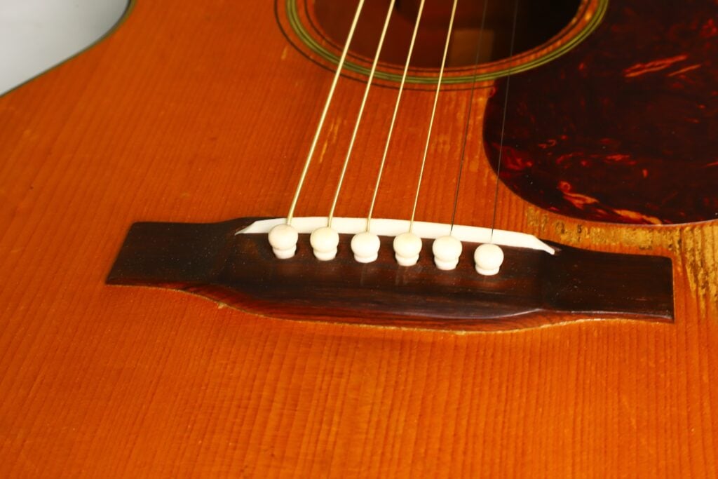 Nahaufnahme des Stegs und der Saiten der Martin 00-18 Akustikgitarre aus dem Jahr 1953, mit sechs weißen Stegstiften auf einem Holzkorpus und einem daneben liegenden Schlagbrett aus Schildpatt. Nahaufnahme des Stegs und der Saiten der Martin 00-18 Akustikgitarre aus dem Jahr 1953, mit sechs weißen Stegstiften auf einem Holzkorpus und einem daneben liegenden Schlagbrett aus Schildpatt.