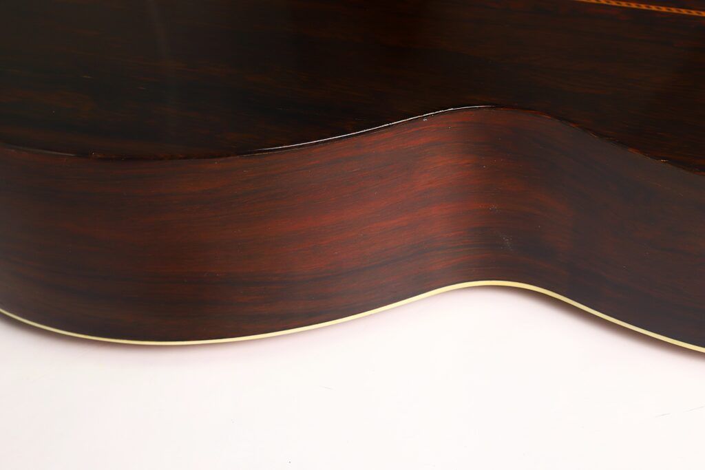Nahaufnahme der Seite der Washburn 1900 Parlor Akustikgitarre mit dunkler, polierter Holzmaserung und einer schmalen, hellen Zierleiste entlang der geschwungenen Kante vor weißem Hintergrund. Nahaufnahme der Seite der Washburn 1900 Parlor Akustikgitarre mit dunkler, polierter Holzmaserung und einer schmalen, hellen Zierleiste entlang der geschwungenen Kante vor weißem Hintergrund.