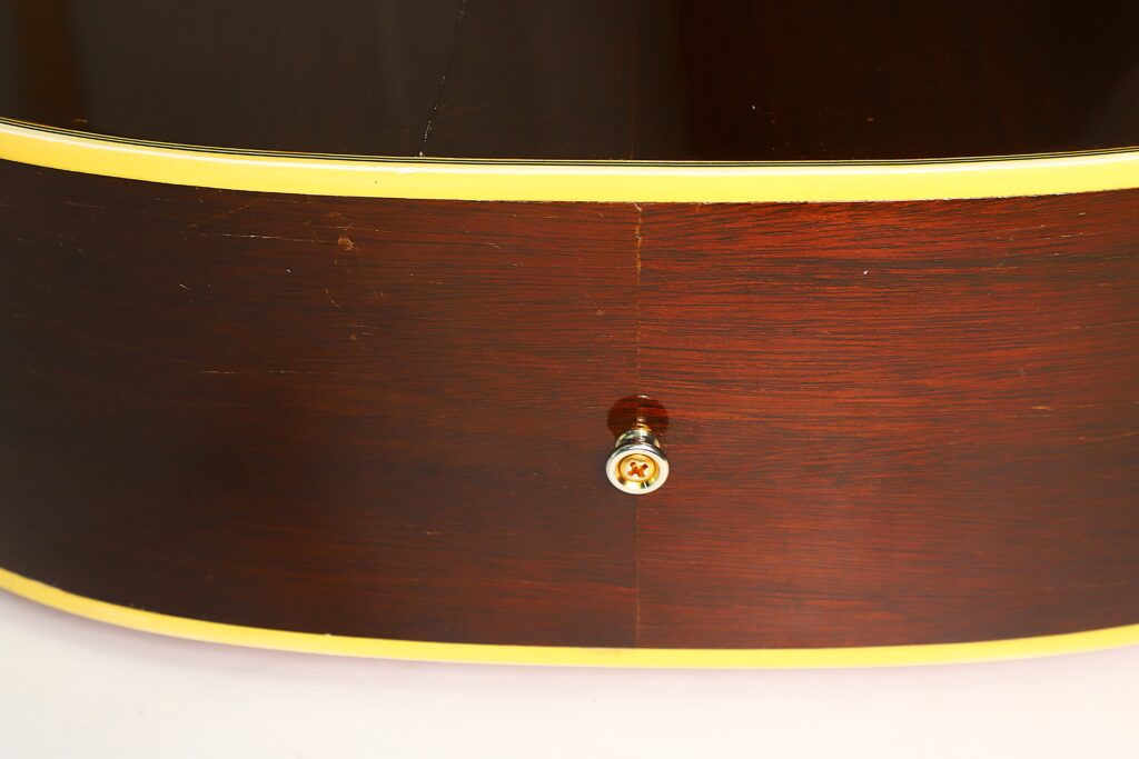 Nahaufnahme der Seite einer Gibson Hummingbird Akustikgitarre aus dem Jahr 1968, die ihre glänzende Holzoberfläche, die gelbe Einfassung und einen Metallgurtknopf in der Mitte nahe einer sichtbaren Naht zeigt. Nahaufnahme der Seite einer Gibson Hummingbird Akustikgitarre aus dem Jahr 1968, die ihre glänzende Holzoberfläche, die gelbe Einfassung und einen Metallgurtknopf in der Mitte nahe einer sichtbaren Naht zeigt.