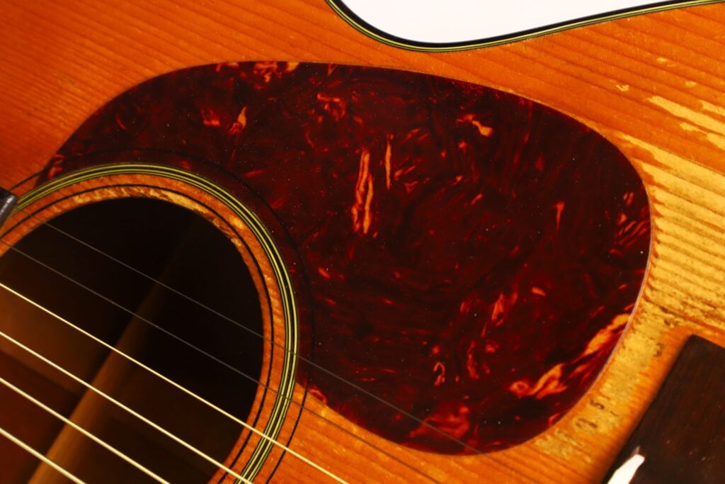 Nahaufnahme einer Martin 00-18 Akustikgitarre aus dem Jahr 1953, bei der das Schallloch, die Saiten und ein marmoriertes, dunkelrot-braunes Schlagbrett auf dem Holzkorpus besonders hervorgehoben sind. Nahaufnahme einer Martin 00-18 Akustikgitarre aus dem Jahr 1953, bei der das Schallloch, die Saiten und ein marmoriertes, dunkelrot-braunes Schlagbrett auf dem Holzkorpus besonders hervorgehoben sind.