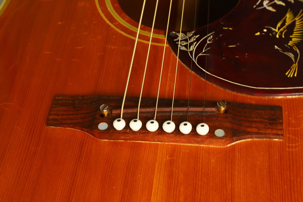 Nahaufnahme des Stegs und der Saiten der Gibson Hummingbird 1968 Akustikgitarre mit Holzkorpus, zu sehen ist das charakteristische, verzierte Schlagbrett mit Vogel- und Blumenmotiv. Nahaufnahme des Stegs und der Saiten der Gibson Hummingbird 1968 Akustikgitarre mit Holzkorpus, zu sehen ist das charakteristische, verzierte Schlagbrett mit Vogel- und Blumenmotiv.