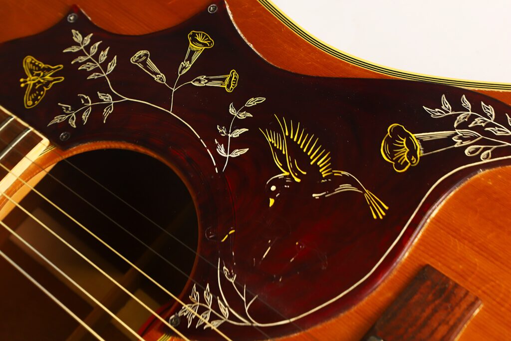 Nahaufnahme einer Gibson Hummingbird Akustikgitarre aus dem Jahr 1968, bei der das dunkle Schlagbrett mit seinen aufwendig eingravierten gelben Blumenmotiven und einem fliegenden Vogel besonders zur Geltung kommt. Nahaufnahme einer Gibson Hummingbird Akustikgitarre aus dem Jahr 1968, bei der das dunkle Schlagbrett mit seinen aufwendig eingravierten gelben Blumenmotiven und einem fliegenden Vogel besonders zur Geltung kommt.