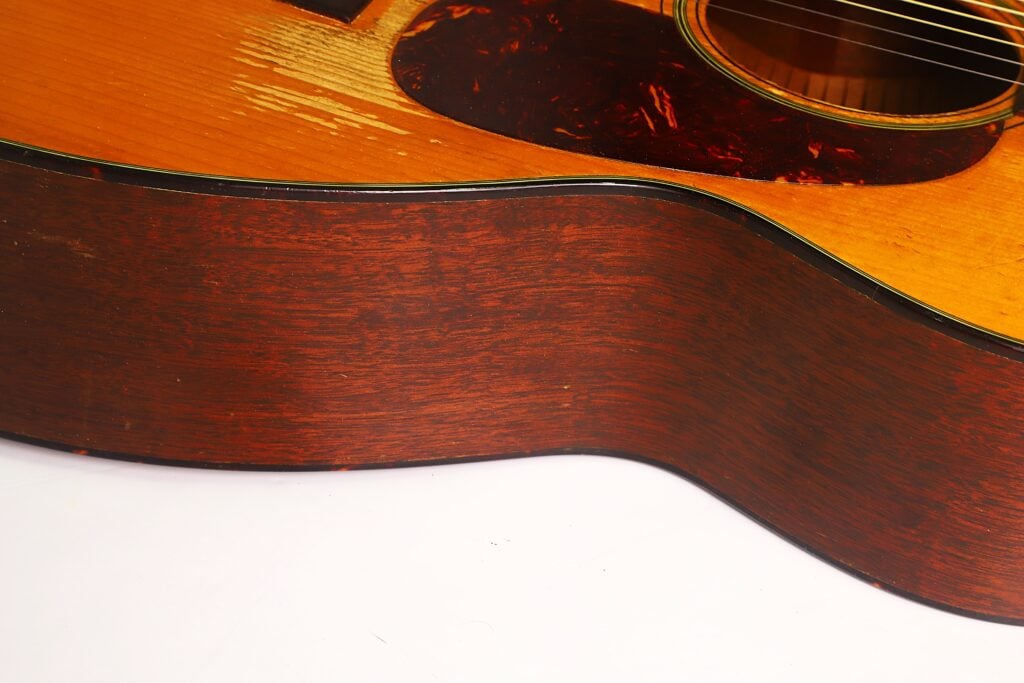 Nahaufnahme der Seite und eines Teils des Korpus einer Martin 00-18 Akustikgitarre aus dem Jahr 1953, die die reichhaltige Holzmaserung, die glänzende Lackierung und einige Saiten zeigt, die oben rechts zu sehen sind. Die Gitarre liegt auf einer weißen Unterlage. Nahaufnahme der Seite und eines Teils des Korpus einer Martin 00-18 Akustikgitarre aus dem Jahr 1953, die die reichhaltige Holzmaserung, die glänzende Lackierung und einige Saiten zeigt, die oben rechts zu sehen sind. Die Gitarre liegt auf einer weißen Unterlage.