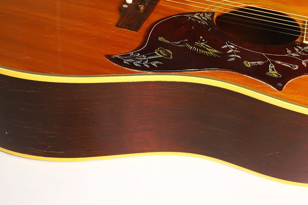 Nahaufnahme des Korpus der Gibson Hummingbird 1968 Akustikgitarre, die die Holzmaserung, die dunkelbraune Seite mit gelber Zierleiste und ein mit Vogel- und Blumenmotiven verziertes Schlagbrett hervorhebt. Nahaufnahme des Korpus der Gibson Hummingbird 1968 Akustikgitarre, die die Holzmaserung, die dunkelbraune Seite mit gelber Zierleiste und ein mit Vogel- und Blumenmotiven verziertes Schlagbrett hervorhebt.