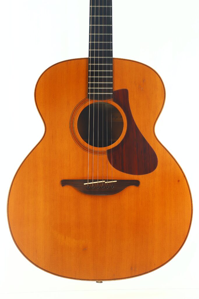 Lowden G-7 Akustikgitarre aus den 1980er Jahren mit naturbelassener Holzoberfläche, dunklem Schlagbrett, sechs Saiten und einem sichtbaren Schallloch, abgebildet vor einem schlichten weißen Hintergrund. Lowden G-7 Akustikgitarre aus den 1980er Jahren mit naturbelassener Holzoberfläche, dunklem Schlagbrett, sechs Saiten und einem sichtbaren Schallloch, abgebildet vor einem schlichten weißen Hintergrund.