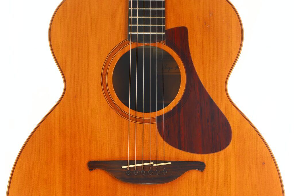 Nahaufnahme des Korpus einer Lowden G-7 Akustikgitarre aus den 1980er Jahren mit sechs Saiten, einem Holzsteg mit weißen Stiften, einem runden Schallloch und einem dunkelbraunen Schlagbrett auf hellem Holz. Der Hintergrund ist weiß. Nahaufnahme des Korpus einer Lowden G-7 Akustikgitarre aus den 1980er Jahren mit sechs Saiten, einem Holzsteg mit weißen Stiften, einem runden Schallloch und einem dunkelbraunen Schlagbrett auf hellem Holz. Der Hintergrund ist weiß.