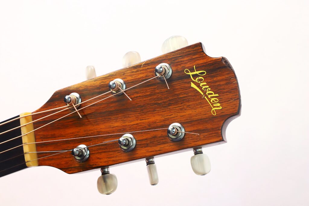 Nahaufnahme des Kopfes einer Lowden G-7 Akustikgitarre aus den 1980er Jahren mit sechs Stimmwirbeln, Saiten und dem Lowden-Logo auf einer Holzmaserung, vor einem schlichten weißen Hintergrund. Nahaufnahme des Kopfes einer Lowden G-7 Akustikgitarre aus den 1980er Jahren mit sechs Stimmwirbeln, Saiten und dem Lowden-Logo auf einer Holzmaserung, vor einem schlichten weißen Hintergrund.
