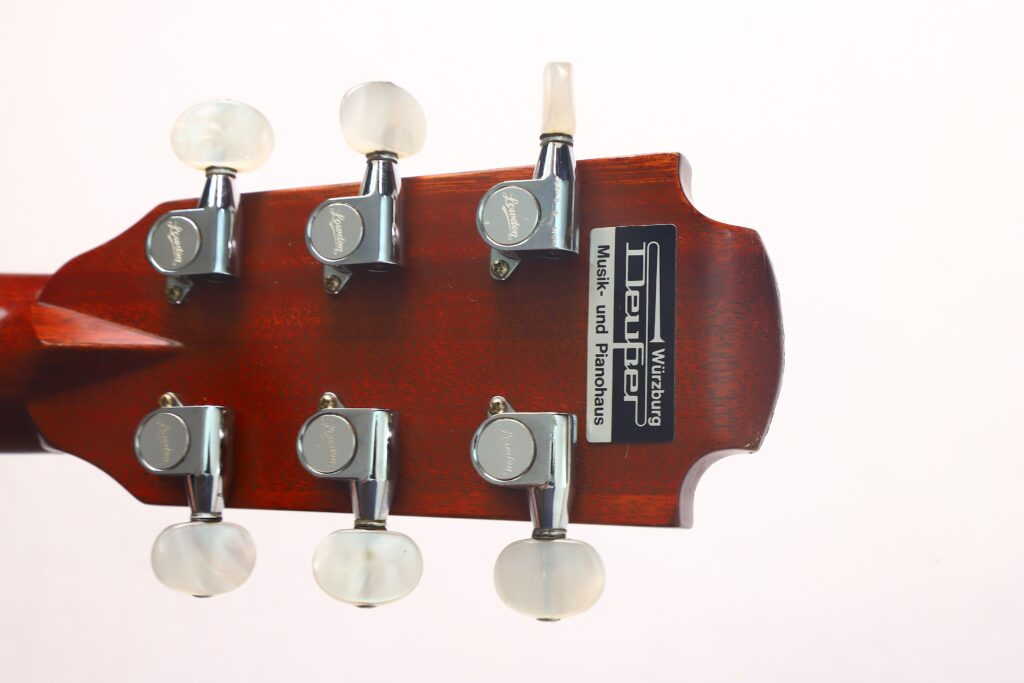 Nahaufnahme der Rückseite des Kopfes einer Lowden G-7 Akustikgitarre aus den 1980er Jahren, auf der sechs silberne Stimmwirbel mit weißen Knöpfen und ein Etikett mit Text zu sehen sind. Die rötliche Holzoberfläche bildet einen Kontrast zum schlichten, hellen Hintergrund. Nahaufnahme der Rückseite des Kopfes einer Lowden G-7 Akustikgitarre aus den 1980er Jahren, auf der sechs silberne Stimmwirbel mit weißen Knöpfen und ein Etikett mit Text zu sehen sind. Die rötliche Holzoberfläche bildet einen Kontrast zum schlichten, hellen Hintergrund.