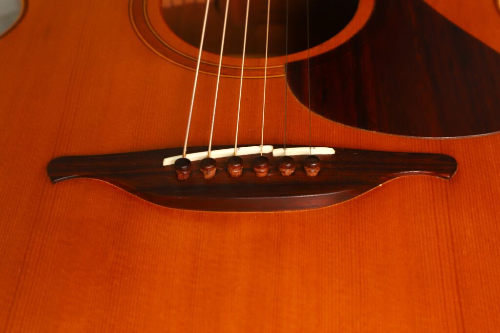 Nahaufnahme des Stegs und der Saiten einer Lowden G-7 Akustikgitarre aus den 1980er Jahren mit Holzkorpus, die feine Details der Holzmaserung und der Einfassung zeigt. Nahaufnahme des Stegs und der Saiten einer Lowden G-7 Akustikgitarre aus den 1980er Jahren mit Holzkorpus, die feine Details der Holzmaserung und der Einfassung zeigt.