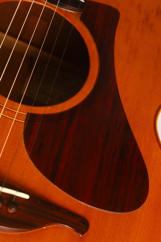 Nahaufnahme des Korpus der Lowden G-7 Akustikgitarre aus den 1980er Jahren, bei der die Saiten, das Schallloch, das dunkle Holz-Schlagbrett und ein Teil des Stegs vor dem Hintergrund der warmen Holzoberfläche hervorgehoben sind. Nahaufnahme des Korpus der Lowden G-7 Akustikgitarre aus den 1980er Jahren, bei der die Saiten, das Schallloch, das dunkle Holz-Schlagbrett und ein Teil des Stegs vor dem Hintergrund der warmen Holzoberfläche hervorgehoben sind.