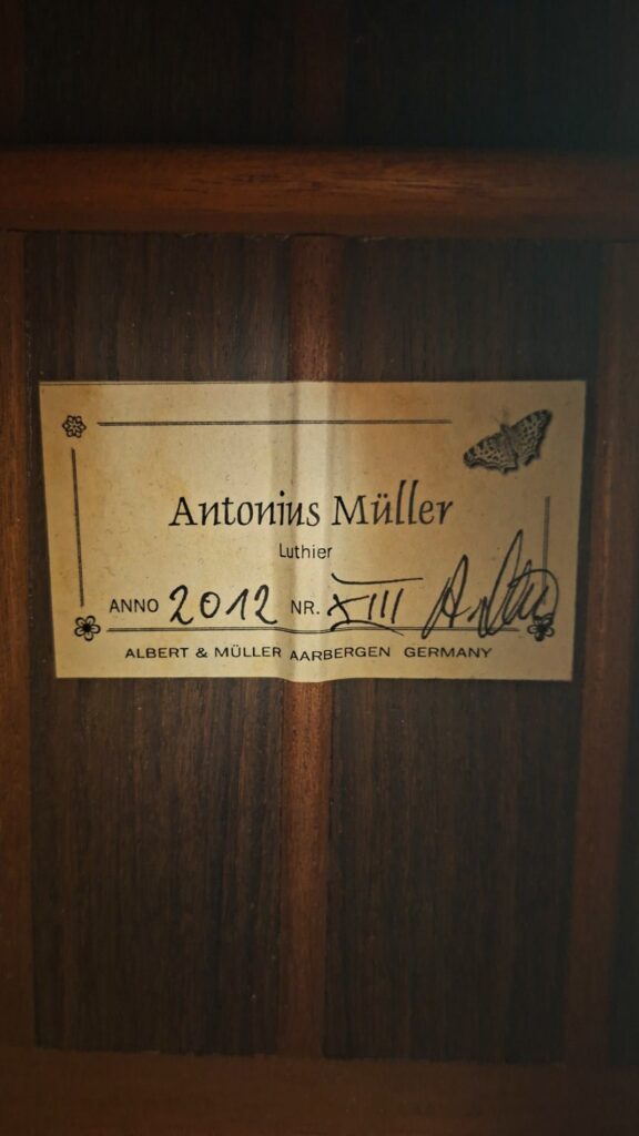 Eine Nahaufnahme des Herstelleretiketts im Inneren der „Antonius Müller 2012 Doubletop Klassische Gitarre“ zeigt den Text „Antonius Müller, Anno 2012, Nr. XIII“ sowie die Abbildung eines Schmetterlings und eine Unterschrift. Darunter steht „Albert & Müller, Aarbergen, Deutschland“. Eine Nahaufnahme des Herstelleretiketts im Inneren der „Antonius Müller 2012 Doubletop Klassische Gitarre“ zeigt den Text „Antonius Müller, Anno 2012, Nr. XIII“ sowie die Abbildung eines Schmetterlings und eine Unterschrift. Darunter steht „Albert & Müller, Aarbergen, Deutschland“.