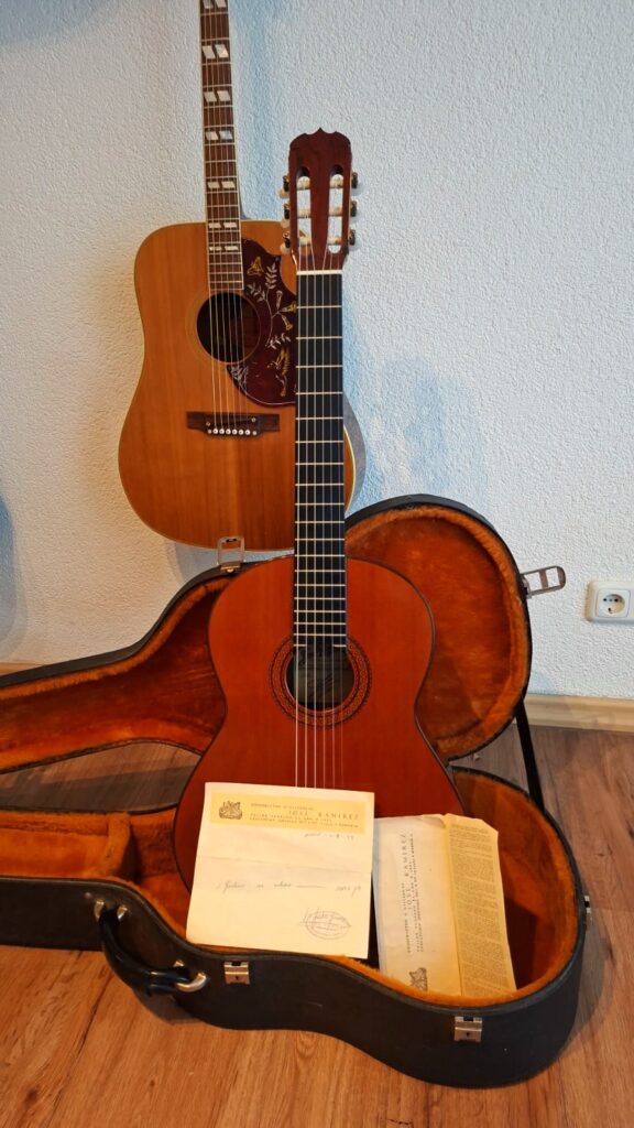 Eine klassische Gitarre von Jose Ramirez, Modell 1a, Baujahr 1977, liegt in einem offenen Koffer mit Unterlagen darin vor einer an der Wand lehnenden Akustikgitarre auf einem Holzboden. Eine klassische Gitarre von Jose Ramirez, Modell 1a, Baujahr 1977, liegt in einem offenen Koffer mit Unterlagen darin vor einer an der Wand lehnenden Akustikgitarre auf einem Holzboden.