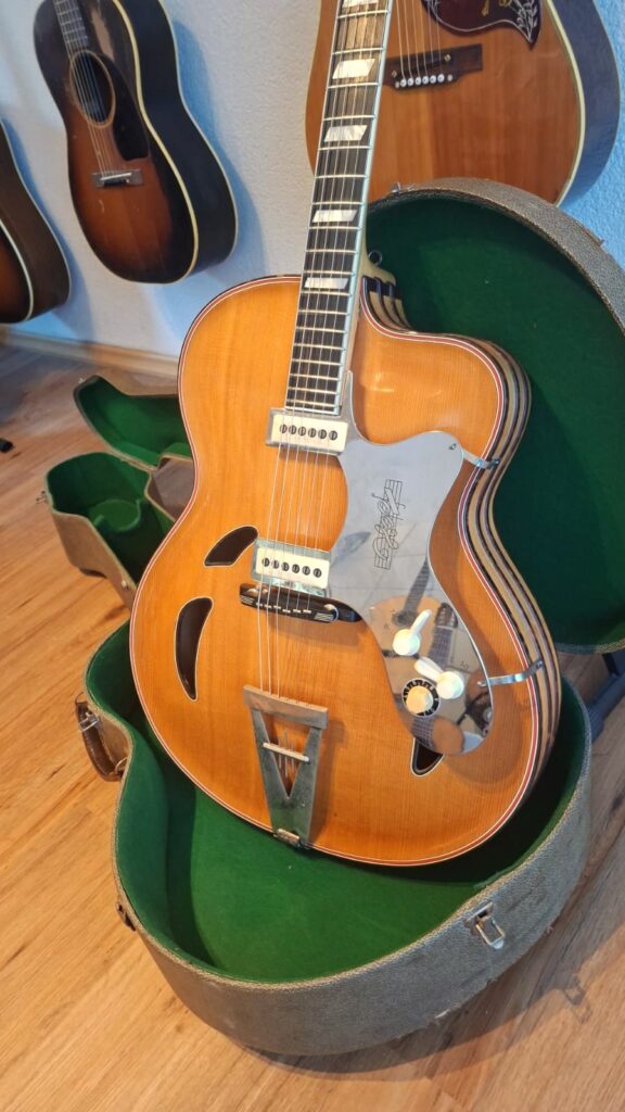 Eine Jazz-Gitarre von Artur Lang aus den 1950er Jahren mit naturbelassener Holzoberfläche und F-Löchern wird in einem offenen, grün ausgekleideten Hartschalenkoffer präsentiert, während mehrere andere Akustikgitarren hinter ihr an der Wand hängen. Die Gitarre verfügt über einen Saitenhalter aus Metall. Eine Jazz-Gitarre von Artur Lang aus den 1950er Jahren mit naturbelassener Holzoberfläche und F-Löchern wird in einem offenen, grün ausgekleideten Hartschalenkoffer präsentiert, während mehrere andere Akustikgitarren hinter ihr an der Wand hängen. Die Gitarre verfügt über einen Saitenhalter aus Metall.