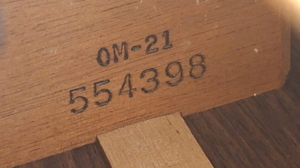 Eine Nahaufnahme einer Holzoberfläche der MARTIN OM-21 Akustikgitarre aus dem Jahr 1995, auf der der Schriftzug „OM-21“ und die Nummer „554398“ in schwarzer Tinte eingeprägt sind. Eine Nahaufnahme einer Holzoberfläche der MARTIN OM-21 Akustikgitarre aus dem Jahr 1995, auf der der Schriftzug „OM-21“ und die Nummer „554398“ in schwarzer Tinte eingeprägt sind.