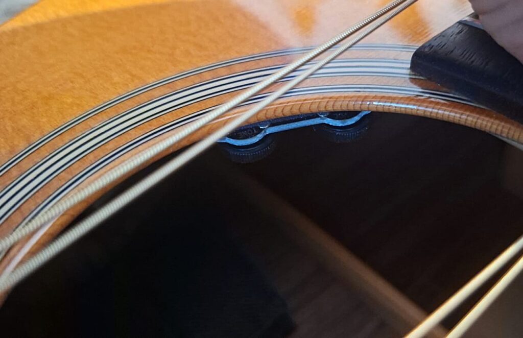 Nahaufnahme der MARTIN OM-21 Akustikgitarre aus dem Jahr 1995, bei der die Saiten und das Schallloch im Vordergrund stehen und die Stegstifte sowie ein Teil des Holzkörpers zu sehen sind. Nahaufnahme der MARTIN OM-21 Akustikgitarre aus dem Jahr 1995, bei der die Saiten und das Schallloch im Vordergrund stehen und die Stegstifte sowie ein Teil des Holzkörpers zu sehen sind.